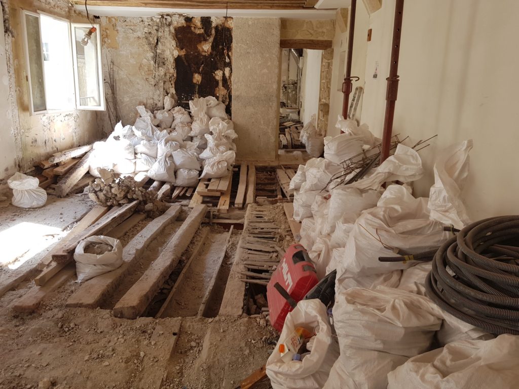 Intérieur de bâtiment en rénovation avec plancher bois partiellement déposé, solives apparentes et grande quantité de sacs de gravats stockés au sol, montrant une surcharge localisée sur une structure ancienne.