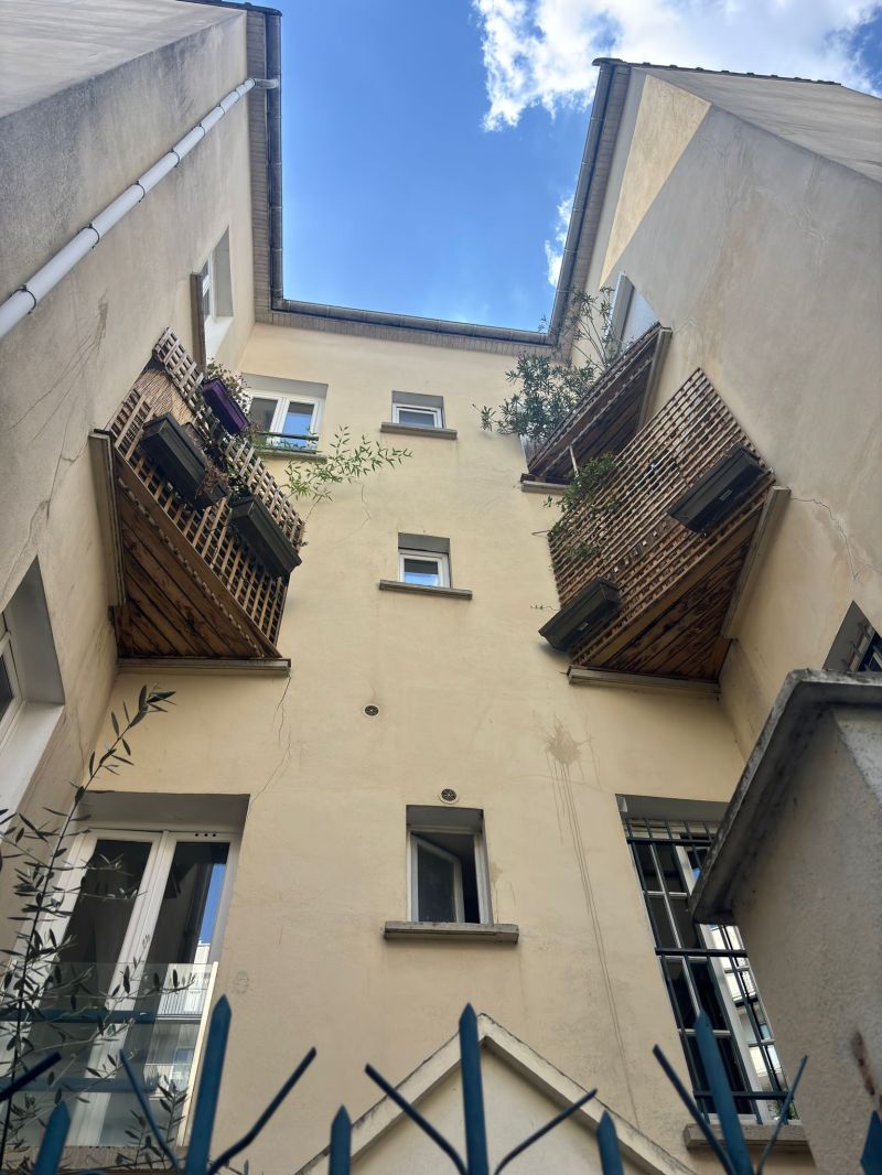 Vue en contre-plongée d'une cour intérieure d'immeuble avec deux petits balcons en bois rapportés, supportant des jardinières. Les balcons semblent reposer sur de simples planches en bois posées sur les appuis de fenêtres, soulevant des interrogations sur leur mode de fixation et leur capacité portante. Des fissures sont également visibles sur les enduits de façade à proximité des balcons.