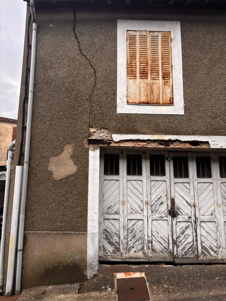 Façade de maison avec une fissure verticale importante partant du haut du mur et traversant l’enduit jusqu’au niveau d’un linteau métallique fortement corrodé au-dessus d’une porte de garage.
